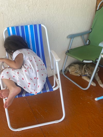 A young child, with dark hair and wearing a white dress with red hearts, is seated on a blue and white striped beach chair, facing away from the camera. Their legs are bare and their feet are visible. To the right of the child's chair is a green folding chair, underneath which a small, light brown puppy is lying down. The puppy appears to be a young dog. Both chairs are situated on an orange-brown wooden floor. The background consists of a light-colored, textured wall. The overall scene suggests an outdoor setting, potentially a patio or porch. The location of Бетюнцы, Russia is not evident from the image itself.