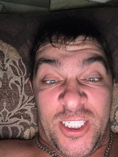 The image is a close-up, overhead shot of a man's face, taken indoors. The man has dark, messy hair, thick eyebrows, and light blue eyes. His face is flushed, and he has a noticeable amount of stubble on his chin and jawline. His mouth is open, revealing white, even teeth, and he is wearing a thick silver chain around his neck. He is lying down, as indicated by the patterned fabric of a cushion or blanket behind him and to his left. The lighting is dim, suggesting it might be nighttime or a dark room. No specific location in Shakhty, Russia is identifiable from the image.