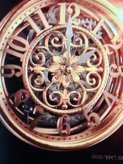 The image shows a close-up of a vintage, ornate pocket watch face. The watch is made of a brass-colored metal with intricate decorative elements. Roman numerals mark the hours, with visible numbers including 10, 11, 12, and 6. The hands of the watch are visible, indicating a time around 11:09. The watch face has a visible gear mechanism beneath the decorative overlay, suggesting it is a mechanical watch. The background is dark and out of focus. In the bottom right corner, there is text that appears to read "OOO 'EPC', O". The location of this object in Cherepovets, Russia, is not discernible from the image itself.