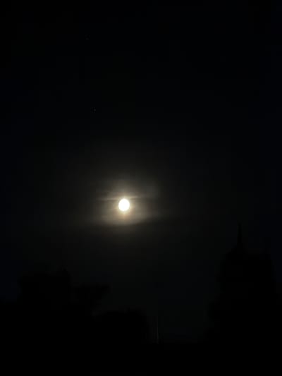 This image depicts a night sky over Khotianivka, Ukraine, with a bright full moon prominently featured in the center. The moon is partially obscured by thin clouds, creating a soft halo effect around it. Two small, faint stars are visible in the upper portion of the sky. Silhouetted against the moonlit sky are dark shapes of trees in the foreground, suggesting a rural or semi-rural setting within Khotianivka. The overall atmosphere is dark and serene, characteristic of a clear night with a bright celestial body.