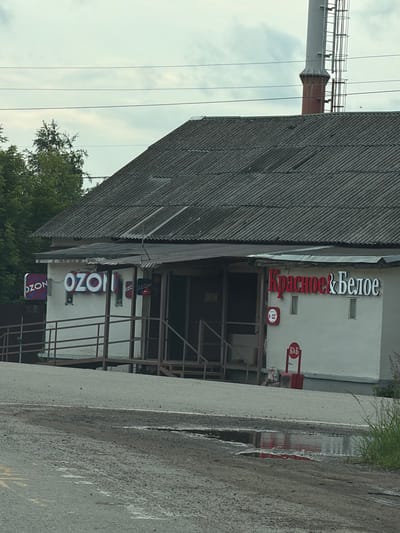 The image shows the exterior of a commercial building, likely a storefront, in Krasnokamsk, Russia. The building has a corrugated metal roof and a white facade. A prominent sign in red and white lettering reads "Красное & Белое" which translates to "Red & White", a common chain of alcohol stores in Russia. Below this, there are signs for "Ozon", a Russian e-commerce company, and a small circular sign that appears to indicate opening hours.
The setting is outdoors, with a paved surface in the foreground, suggesting a road or parking area, with a puddle reflecting the building. Overcast skies are visible, indicating a cloudy day. To the left of the building, a tall industrial structure, possibly a chimney or part of a larger complex, is visible. Trees and greenery are present around the building, suggesting a semi-urban or industrial fringe location. The overall impression is one of a functional commercial area, likely during daylight hours. There are no people visible in the image.