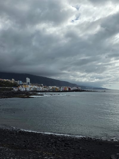 The image shows a coastal view of Puerto de la Cruz, Spain, on a cloudy day. The foreground is a black pebble beach with gentle waves lapping at the shore. The sea is a deep gray, with a few small whitecaps visible. In the midground, a line of colorful buildings, typical of the Canary Islands, stretches along the coastline. Behind the town, a steep, dark mountain range rises, partially obscured by heavy clouds. Some people are visible in the water, swimming or wading near the shore. The overall atmosphere is subdued, with the overcast sky casting a muted light on the scene.