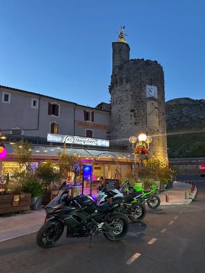 The image depicts a twilight scene in front of the restaurant "Les Templiers" in Anduze, France. A stone tower, possibly a historical landmark, stands to the right of the restaurant building. The tower features a clock and a sundial. Several motorcycles are parked on the street in front of the restaurant. The restaurant has an illuminated sign and outdoor seating areas with plants and lights. A street lamp with decorative elements is also visible near the tower. The sky is a deep blue, indicating the late hour.