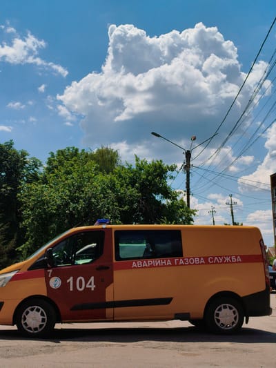 In Velyka Bahachka, Ukraine, a yellow and orange service van with the text "АВАРІЙНА ГАЗОВА СЛУЖБА" (Emergency Gas Service) printed on its side is parked on a street. The van has a blue flashing light on its roof. The side of the van also displays the number "104" and a circular logo. The vehicle is positioned on a paved surface with shadows cast by the sun. Behind the van, a line of green trees is visible under a bright blue sky with large, white cumulus clouds. Utility poles with a complex network of wires are also prominent features of the environment. The scene appears to be set during daylight hours.