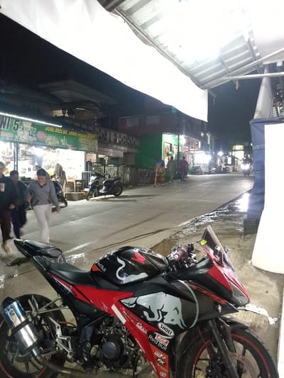 The image depicts an evening street scene in Caringin, Indonesia, specifically near "JL. RAYA PASAR CIKERETEG KM. 19 BOGOR," as indicated on a shop sign. The sky is dark, suggesting nighttime, with artificial lighting from streetlights and commercial establishments illuminating the area.
In the foreground, a black and red Honda CBR sport motorcycle is parked on a paved surface on the right side of the frame, angled towards the left. The motorcycle features "Honda," "Red Bull," and "Shoei" branding, a chrome exhaust, and red accents on its wheels and bodywork. Adjacent to the motorcycle on the far right, a partial blue tarp structure is visible.
To the left of the motorcycle, a paved road stretches into the distance. Several wet patches are visible on the road surface. Approximately five individuals are walking away from the camera on the left side of the road. One person in a gray top and light-colored trousers is centrally positioned within this group.
A row of commercial buildings lines the left side of the street. One prominent shop features a green sign with white text, partially visible as "UNIT 5," "JUAL BELI SR. UHAN SELAR," and "FROZEN FOOD." A dark scooter is parked near this shop. Further down the street, additional pedestrians are visible, along with multiple illuminated vendor stalls or small businesses. Another motorcycle with a rider is present in the distant background on the right, identifiable by its illuminated headlight. Overhead, a white tarp or awning provides cover in the foreground.