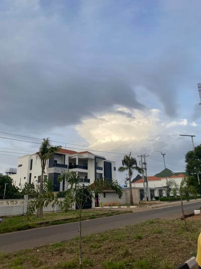 This image captures a scene in Barikin Jirga Summa, Nigeria, on a day with dramatic cloud formations dominating the sky. In the foreground, a paved road runs horizontally across the frame, bordered by grassy verges with scattered dry patches and young trees. On the left side of the road, a modern, multi-story white building with dark window accents and orange roof trim stands prominently, partially obscured by a tall palm tree. A low, textured white wall with a black metal fence separates the building from the road. Further down the road, another building with a green roof and orange trim is visible. The sky is filled with a mix of dark grey and bright white clouds, suggesting changing weather conditions. Power lines and utility poles are visible, crossing the sky above the buildings. Two individuals, one wearing pink and the other yellow, are standing near the entrance of the building on the left. The overall impression is of a residential or commercial area under an overcast sky.