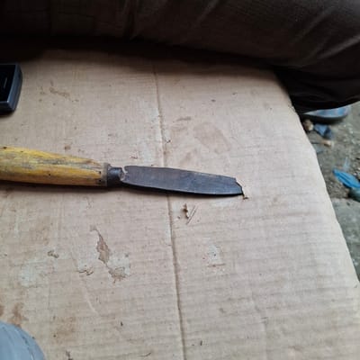 The image depicts a close-up view of a broken knife lying on a piece of cardboard. The knife has a yellow wooden handle and a rusty metal blade that appears to be chipped and worn. The cardboard surface is textured and shows some signs of wear and tear, with a few stains and indentations. In the background, there is a dark, textured surface, possibly fabric or upholstery, and a hint of what appears to be a person's leg or clothing. The environment appears to be outdoors or in a dimly lit indoor space, with a rough, possibly unpaved ground visible in the lower right corner. There are no explicit indicators of the location being Mesjid Tuha, Indonesia, beyond the context provided. The scene is static, with the knife simply resting on the cardboard.
