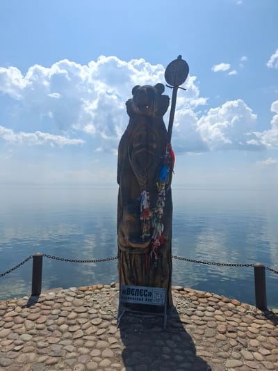 This image depicts a wooden sculpture of a bear adorned with colorful ribbons. The sculpture is positioned on a cobblestone surface overlooking a body of water. A chain fence runs along the edge of the cobblestone path, separating it from the water. In the background, a vast expanse of water stretches to the horizon under a bright, cloudy sky. The lighting suggests it is daytime, likely late morning or early afternoon, with the sun casting shadows from the sculpture and the fence posts. The clouds are white and fluffy, indicating fair weather.

A sign with Cyrillic text is visible near the base of the sculpture. The text reads "«ВЕЛЕСТ» Славянский Бог" which translates to "«VELES» Slavic God." This suggests the sculpture represents the Slavic deity Veles, often depicted as a bear or having bear-like attributes. The presence of the ribbons tied to the sculpture is a common folk tradition, often associated with wishes or prayers.

The overall scene evokes a sense of cultural heritage and a connection to nature, given the representation of a bear and the expansive natural landscape. The location is identified as Listvyanka, Russia, a settlement on the shores of Lake Baikal, which aligns with the visual cues of a large body of water and the surrounding natural environment.