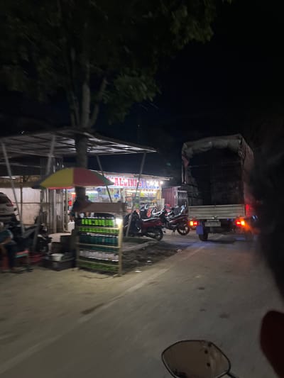 The image depicts a roadside scene at night in Bundar, Indonesia. A small open-air market is visible on the left, with a colorful umbrella providing shade. Shelves displaying rows of green bottles, likely beverages, are prominently featured. A few motorcycles are parked nearby. In the background, a large truck is parked on the right side of the road, with its taillights illuminated. The overall environment suggests a commercial area operating into the evening.