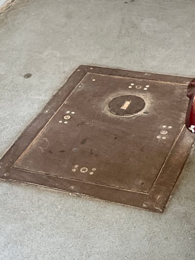 The image shows a weathered, rectangular metal plate set into a concrete surface. The plate is dark brown and appears to be made of iron or a similar metal, exhibiting signs of rust and wear. It has a raised border around its perimeter, secured by small rivets at the corners. Centered on the plate is a circular indentation with a rectangular slot in its middle. Decorative patterns, consisting of small white dots arranged in groups of three, are visible on the plate at various points. The concrete surface surrounding the metal plate is a light gray color and shows a few small debris. Part of a dark red, cylindrical object with white accents is visible on the right side of the image, partially obscuring the metal plate. There is no indication of the location within the city of Hlevakha, Ukraine, in the image.