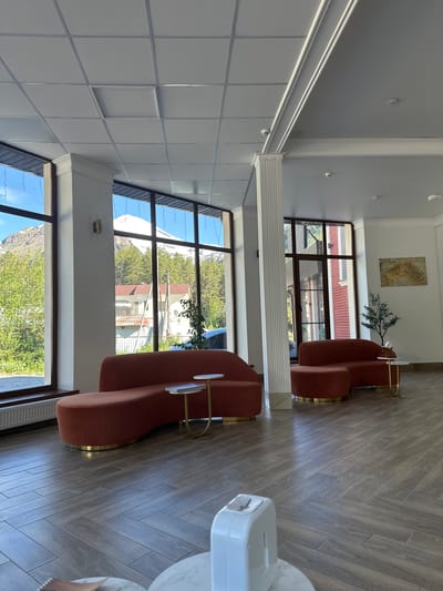 The image depicts a well-lit, spacious interior that appears to be a hotel lobby or a common area in a building in Terskol, Russia. The room features large windows offering a view of snow-capped mountains, evergreen trees, and residential buildings, suggesting a mountainous resort town.

Dominating the foreground are two plush, rust-colored curved sofas with gold-colored bases. Between them, small, modern side tables with white tops and gold legs are placed. One table has a stack of books on it, and the other has a small plant. In the middle of the room, a white dispenser, possibly for hand sanitizer, sits on a pedestal with a marble-patterned top. The floor is laid with light-colored, wide wooden planks arranged in a herringbone pattern.

The ceiling is a dropped ceiling with a grid of white acoustic tiles and some recessed lighting fixtures. Ornate white molding frames the upper parts of the walls and columns. On the far wall, a framed abstract artwork with golden hues hangs above a small potted olive tree.

The overall impression is one of modern comfort and elegance, with a clear connection to the natural mountain landscape visible through the windows. There are no people visible in the image.
