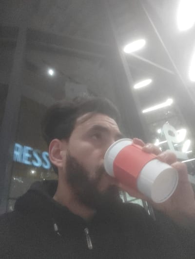 A bearded man in a black hoodie is drinking from a white cup with a red sleeve. The background is blurred, but a sign with the letters "RESS" is visible. Above, there are several long, bright lights in the ceiling. The man is looking slightly upwards and to the right. The photo is taken at an angle, looking up at the man.