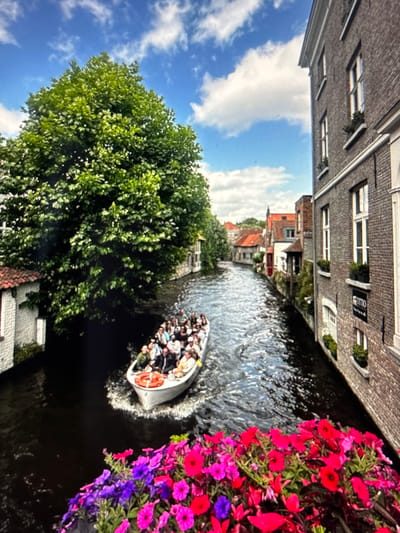 The image shows a canal scene with a boat filled with people. The boat is white and appears to be a tour boat, with many passengers looking around. The canal is surrounded by buildings with brick facades and windows. There is a lush green tree on the left side of the image, and a prominent display of colorful flowers, predominantly pink and purple, on the right. The water in the canal is dark and reflects the surrounding environment. The sky is blue with scattered white clouds. The overall impression is a picturesque, possibly tourist-oriented scene, suggesting it is a pleasant day. The image is oriented vertically, with the top of the buildings appearing at the bottom of the frame. There is some visible text on a sign on one of the buildings, but it is too blurry to read. The provided location context of Pastavy, Belarus, suggests this is a canal within that city.