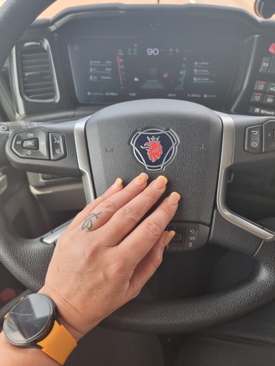 A person's right hand, adorned with pale yellow-tipped fingernails and a small black tattoo resembling a stylized leaf on the index finger, rests on the black, textured steering wheel of a Scania truck. The center of the steering wheel displays the Scania griffin logo. A smartwatch with a circular, cracked black screen and a vibrant orange band is visible on the individual's wrist. The vehicle's digital dashboard is visible through the steering wheel, displaying "90" (likely kilometers per hour) prominently, along with other vehicle information and indicators, including a "90 km/h" label on a steering wheel control. The scene is located within the cabin of a heavy goods vehicle in Villafranca del Campo, Spain.