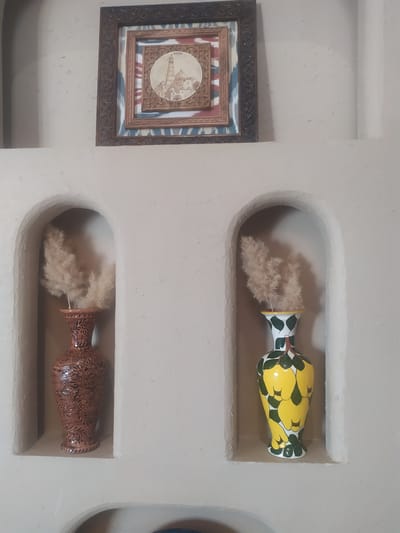 The image shows an interior scene with decorative niches built into a light-colored wall. A framed artwork hangs above the niches. The artwork features a detailed depiction of a city with a tall minaret, identified by the text "KHIYA" (Khiva) at the top. The artwork is surrounded by layers of decorative frames, including one with a geometric pattern and another with blue and red patterned fabric.

Below the artwork, two arched niches are visible. The left niche contains a terracotta-colored vase with intricate brown carvings and dried decorative reeds. The right niche holds a yellow vase adorned with a green leaf and fruit pattern, also containing dried decorative reeds. The overall impression is of a tastefully decorated interior space, possibly within a traditional building in Khiva, Uzbekistan, given the textual reference in the artwork.