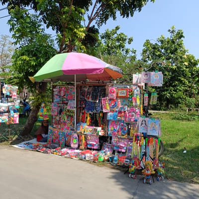 This is a full-shot, eye-level, outdoor photograph of a toy stall in Kebondalem, Indonesia. The stall is set up on the side of a paved road, with a grassy area to the right and trees and foliage in the background. The stall is covered by a multi-colored umbrella, with sections of pink, green, and orange. A vendor, wearing a brown hijab and a red shirt, sits on a white cloth at the left side of the stall, partially obscured by the display.

The stall is densely packed with a variety of toys, primarily packaged children's toys. On display are dolls, toy vehicles (including cars and trucks), toy guns, toy kitchen sets, and other playthings, mostly in bright colors. The toys are arranged on shelves, hanging displays, and spread out on the ground in front of the stall. A blue motorcycle is parked behind the stall. The environment is sunny with clear blue skies, and the trees indicate a warm climate. The overall scene depicts a typical street-side vendor in Indonesia, showcasing a wide assortment of toys for sale.