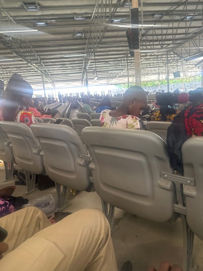 A large gathering is observed within a spacious, covered structure in Ozuaha, Nigeria. The structure features a corrugated metal roof supported by an exposed steel truss system and a visible wooden support column with an attached speaker. Rows of light grey, plastic stadium-style seating fill the area. In the immediate foreground, the lower legs of an individual wearing tan trousers and holding a dark smartphone are visible. To the left, a person's bare feet with red-painted toenails are present. Further into the mid-ground, a young child with a shaved head and a gold hoop earring, wearing a light floral dress, is seated and looking downwards. To the child's left, a woman with dark hair, wearing a vibrant patterned top, is seated with a white smartphone placed on the seat beside her. Numerous other individuals are seated in the background, occupying many of the rows, generally facing away from the camera. Objects on the floor include plastic bags and other small personal items. The environment is brightly lit, suggesting daylight.
