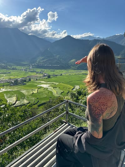 A person, seen from behind and the right side, is seated on a metal slatted bench on a viewing platform, observing a valley in Tú Lệ, Vietnam. The individual has long brown hair and prominent floral/geometric tattoos in red, orange, and green on their right shoulder blade and upper arm. They are wearing a dark grey tank top and a dark hooded jacket over black pants, with a mobile phone resting on their lap. Their right hand is raised, shielding their eyes from the bright sunlight. The panoramic view showcases a wide valley filled with vibrant green, irregularly shaped, and partially submerged rice paddies, indicating terraced cultivation. A winding river flows through the valley, dotted with small settlements and roads. In the background, multiple layers of dark mountains rise under a bright blue sky with scattered white cumulus clouds, from which visible sunbeams descend onto the mountain slopes. A metal railing is visible in the lower right and foreground of the platform.