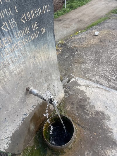 The image shows a water fountain in Lehvaz, Armenia. The fountain is made of gray stone with inscriptions in a foreign language. A metal spout dispenses water into a black metal basin. The surrounding ground is concrete and gravel. A path borders the fountain. No people are visible.