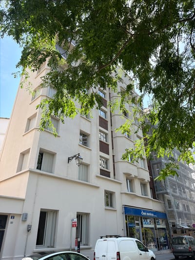 The image shows a narrow street in Courbevoie, France, with a multi-story building and a smaller building under construction. The multi-story building features a stucco facade with several windows and balconies. A tree with lush green leaves partially obstructs the view of the building, casting dappled sunlight onto the scene. The smaller building to the right is covered in scaffolding and white netting, indicating ongoing construction or renovation. On the ground floor of the multi-story building, there is a storefront with a blue awning and the text "Omni medical". Two vehicles, a white van and a silver car, are parked on the street in front of the buildings. The sky is clear and blue.