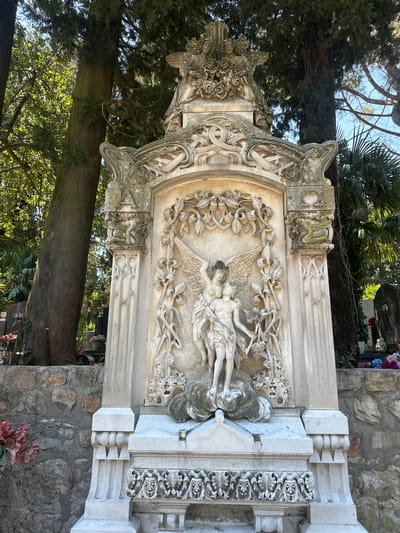 This is a vertical, eye-level, outdoor, medium shot of an ornate, weathered stone monument in a cemetery. The monument is intricately carved with floral motifs, swirling patterns, and a central relief of two winged figures embracing. The figures are rendered in a classical style, with drapery and flowing hair. One figure appears to be an angel, with large feathered wings. The monument is made of light-colored stone and shows signs of age and weathering, with some green moss and lichen growth.
The monument is set against a backdrop of lush green trees and a bright blue sky, suggesting a sunny day. In the background, other headstones and a stone wall are visible, indicating the cemetery setting. Some artificial flowers are placed at the base of the monument and on a nearby grave. The overall mood is one of solemnity and artistic appreciation, with the detailed carvings suggesting a memorial of significance. There is a visible numerical inscription "607" on a dark plaque in the lower right corner, but no other legible text is discernible. The light suggests it is daytime.
