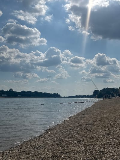 This media file depicts a serene outdoor landscape, appearing rotated 90 degrees counter-clockwise. The scene captures a bright, sunny day over a body of water with a pebble shore, situated in the Cukarica Urban Municipality, Serbia.
The sky, visible in the upper part of the correctly oriented image, is a clear blue, adorned with numerous large, white cumulus clouds. The sun, positioned in the upper left quadrant, emits a strong glare and prominent sunrays that pierce through the clouds.
Below the sky, a calm expanse of water, likely a lake or river, gently shimmers under the sunlight. Several small, red and white buoys are visible floating in the water, suggesting a designated area for swimming or other water activities. A small, light-colored object, possibly another buoy or debris, is also seen floating further out.
Adjacent to the water, a wide and extensive pebble beach stretches across the right side of the frame. The shore is covered uniformly with small, dark-colored pebbles or gravel, creating a distinct texture.
In the distance, beyond the water, a dense line of dark green trees forms a natural horizon. Above this treeline, a tall, white or light-colored crane structure is prominently visible, indicating human development or construction activity in the background.
The image conveys a sense of tranquility and openness, characteristic of a pleasant day at a natural waterfront location. The time of day appears to be midday or early afternoon due to the high sun and bright light. No people or discernible text are visible in the image.
