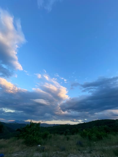 The media file shows a landscape scene with a bright blue sky and dramatic clouds. The clouds are a mix of dark and light, with some illuminated by sunlight, suggesting either sunrise or sunset. The sky takes up the majority of the frame, with a band of dark green trees and rolling hills on the right side, indicative of a mountainous or hilly terrain. The vegetation appears lush and natural, suggesting a rural or wilderness setting. The lighting and cloud formations create a sense of depth and atmosphere. There are no people or specific objects clearly visible in the image, nor any discernible text. The overall mood is one of natural beauty and perhaps a touch of drama due to the cloudy sky. The location context of Ablanitsa, Bulgaria, places this scene in a European natural environment, likely a rural area with significant elevation changes.