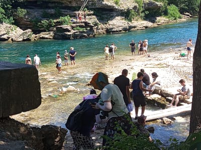 The image depicts a scenic view of the Bližanjka River in Mostar, Bosnia and Herzegovina, a popular natural attraction known for its clear turquoise waters and rocky surroundings. A group of people are engaged in various activities along the riverbank. Some are wading in the shallow water, while others are standing on the rocky shore. In the foreground, several individuals are dressed casually, some with backpacks, suggesting a day trip or recreational outing. One person in the center is wearing a headscarf and holding a smartphone. Further back, a group of people are gathered near the water's edge, with some appearing to be preparing to enter the river. In the background, a steep, rocky cliff face is visible, covered in lush green vegetation, with a wooden ladder leading to an elevated platform where a few individuals are standing. The river flows gently, with sunlight illuminating the clear water and the pebbles on the riverbed. The overall atmosphere is one of leisure and enjoyment of the natural environment.