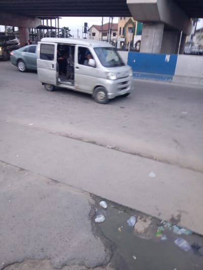 The image depicts a street scene in Port-Harcourt, Nigeria, likely under an overpass or bridge. In the foreground, a silver minivan with its passenger side door open is positioned on the asphalt road. Several passengers are visible inside the minivan, some appearing to be in motion or interacting. To the left of the minivan, a light green sedan is stopped, with its front tire turned outwards. Behind the sedan, another vehicle is partially visible. The road surface shows wear and tear, with a visible drain or ditch running along the edge of the road in the lower right portion of the image, containing some debris and murky water. In the background, buildings and structures are visible, along with overhead infrastructure, suggesting an urban environment. The sky is overcast, indicating potentially cloudy weather.