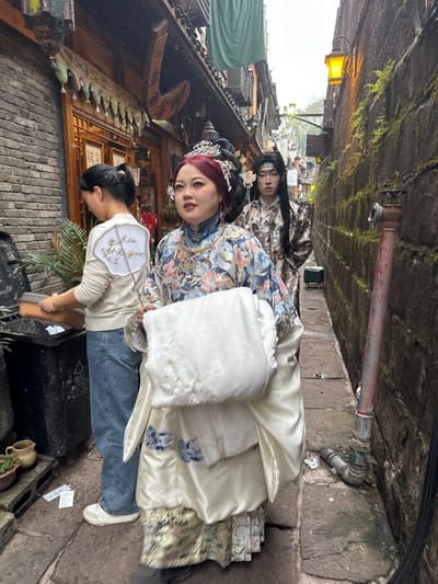 The image depicts a narrow, damp alleyway in the historic city of Fenghuang, China. Three individuals are prominently featured, with two dressed in traditional Chinese attire.

In the foreground, a woman faces slightly right of the camera, wearing an elaborate traditional Chinese costume. Her hair is styled in an ornate black updo with red accents, adorned with a large, multi-jeweled phoenix crown and hairpins. She wears a high-collared robe with a blue, pink, and white floral pattern, over a lighter, possibly satin, undergarment that extends to the floor. She holds a folded white garment with subtle white embroidery. Her expression is neutral.

Behind her, to the left, another woman with dark hair tied in a ponytail is seen from the back, facing a black, open-lidded bin. She wears a cream-colored long-sleeved top, blue jeans, and white sneakers. She holds an orange rectangular tray over the bin, appearing to be disposing of or organizing contents. A white circular fan with green and yellow floral embroidery is slung over her left shoulder.

Further down the alley, behind the first woman, a third individual, likely male, is visible in traditional Chinese attire. They wear a patterned dark robe and have long dark hair with a simple dark headpiece. Their gaze is directed forward.

The alley features a brick wall on the left, leading to traditional wooden buildings with ornate facades, windows, and hanging decorations, including a green fabric banner and a carved wooden sign. Potted plants and discarded papers are on the ground near the black bin. On the right, a tall, narrow brick wall is covered with patches of green moss and ferns. A yellow-lit antique-style lantern is mounted high on this wall. A red vertical pipe with a metal fitting runs along the right wall. The ground consists of irregular stone paving, appearing damp. The sky overhead is overcast. In the distant background, more traditional buildings and a small green cart are visible.