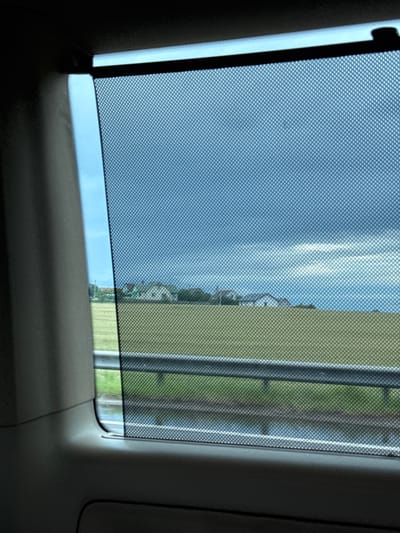The image shows a view through a car window with a mesh sunshade, looking out onto a rural landscape. The sky is overcast with dark clouds, suggesting impending rain or a stormy day. Below the sky is a strip of green field, and further down is a metal guardrail. Beyond the guardrail, a wet surface, possibly a road or pavement, reflects the sky. In the distance, several houses with red and green roofs are visible amidst trees, characteristic of the rural settlements in the Фаніпальскі сельскі Савет region of Belarus. The foreground is dominated by the interior of the car, with a dark gray trim on the left and the sunshade obscuring part of the view.