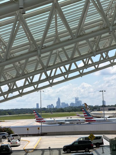 This media file shows an airport scene in Charlotte, USA. The foreground features a large, modern architectural structure with a white metal beam framework and a partially visible glass roof. In the background, an American Airlines plane is parked at the gate, its tail with the iconic red, white, and blue stripes prominently displayed. The plane has the American Airlines logo on its fuselage. Beyond the tarmac, a city skyline with numerous buildings can be seen in the distance, set against a backdrop of trees. Several vehicles, including vans and cars, are parked near the terminal building. The sky is partly cloudy, suggesting a pleasant day. The overall scene depicts a typical airport operational environment. No people are clearly visible. The time of day appears to be daytime, likely late morning or afternoon, given the lighting.