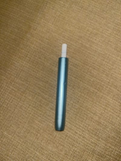 A light blue, cylindrical electronic device, approximately 9 cm in length, is vertically centered on a light brown, woven fabric surface. The device features a white, cylindrical tip extending from its upper end, exhibiting a faint, light blue, curved line pattern. The background fabric displays a consistent cross-hatch texture. No individuals, actions, or interactions are depicted. The location is Pedrouços, Portugal.