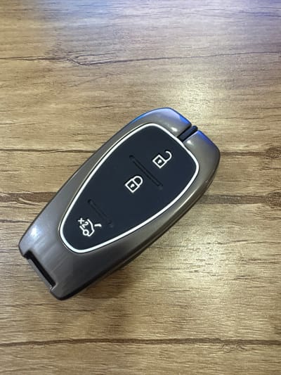A dark grey car key fob with a black insert is centered on a light brown wood-grain surface. The fob has three white icons: an unlocked padlock, a locked padlock, and a car outline with "x2" underneath, suggesting a remote start or trunk feature. The black insert is surrounded by a thin white border, adding a subtle contrast to the dark grey casing. The key fob has a sleek, modern design.