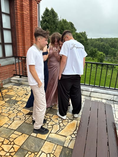 In Zavod Mikhailovskii, Russia, four young adults, two males and two females, are gathered on a tiled balcony overlooking a wooded area. The male on the left wears a white polo shirt and light-colored pants. To his right, a female in a dark dress stands next to another female in a long, shimmering rose-colored dress, who is looking at a smartphone. A second male on the right wears a white t-shirt with black text and black pants. A wooden table is positioned in the foreground. The sky is overcast, and trees are visible in the background.