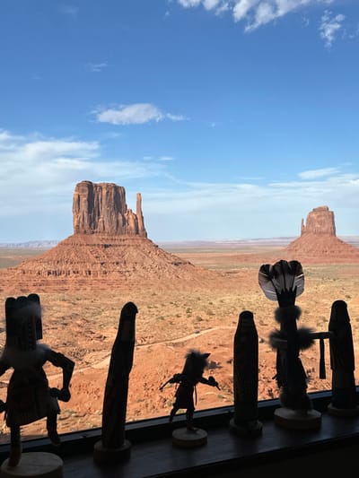 The image shows a view of Monument Valley, a Navajo Nation territory in Oljato-Monument Valley, United States, from what appears to be an indoor vantage point. In the foreground, a row of several Kachina dolls are lined up on a dark surface, silhouetted against the bright outdoor scenery. These dolls are intricately carved and decorated with feathers and fur. The backdrop is dominated by the iconic red rock formations of Monument Valley, with Mittens and Merrick Butte clearly visible. The landscape is arid, with sparse vegetation and a winding road visible in the distance. The sky is a vibrant blue with scattered white clouds, suggesting a clear day.