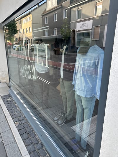 The image captures a storefront window display in the city of Waldbröl, Germany. The reflection on the glass reveals a street scene with buildings, parked cars, and a pedestrian walkway. Inside the store, mannequins are displayed. One mannequin is dressed in a light blue long-sleeved shirt and light-wash jeans, with athletic shoes. Another mannequin appears to be wearing similar attire, partially obscured by the first. Large white lettering on the window reads "RAULI MENSWEAR". The street in front of the store is paved with cobblestones. The store signage above the entrance on the building across the street is visible and appears to read "C. BRAUN". The time of day suggests it is either morning or late afternoon due to the angle of the sunlight and the shadows.