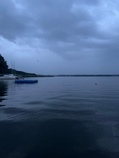 A wide shot captures a lake in Schwielowsee, Germany, under a cloudy, twilight sky. In the foreground, dark, rippling water fills the bottom half of the frame. In the mid-ground, a blue floating dock with a metal ladder is positioned on the left. Beyond the dock, a white sailboat is moored. The shoreline, lined with trees, forms a dark line across the horizon. Orange buoys are visible scattered across the water. The overall atmosphere is dim and moody, suggesting impending or recent rain.