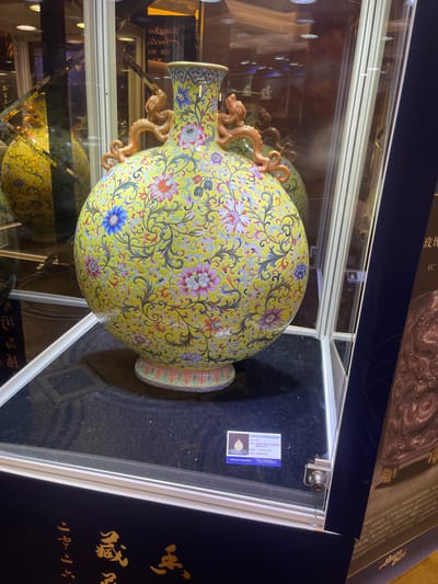 A large, round, yellow ceramic vase with ornate floral and dragon designs is displayed in a glass case. The vase has two dragon-shaped handles on its upper body and is decorated with pink, blue, and red flowers. The background shows reflections of a retail environment, including display shelves and lighting fixtures. The vase is placed on a black velvet stand within the glass enclosure. A small white card with Chinese text and what appears to be an auction lot number is affixed to the inside of the glass case, near the bottom. The overall setting suggests an exhibition or retail display, likely within a commercial establishment. The text on the card and the signage visible in the reflections suggest the location is Futian District, China, possibly within an auction house or luxury goods store.