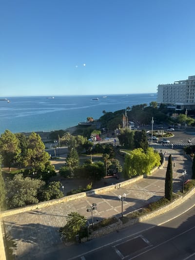 The image depicts a high-angle view of a coastal area in Tarragona, Spain. The Mediterranean Sea dominates the background with several cargo ships visible in the distance. A tree-lined promenade runs along the coast, bordered by a park with various trees and greenery. To the right, a multi-story hotel building stands, adjacent to a roundabout with circulating traffic. The foreground features a paved terrace or walkway with ornate lampposts and low stone walls. Shadows cast across the paving suggest it is mid-morning. A few people are walking on the sidewalk. The sky is clear and blue.