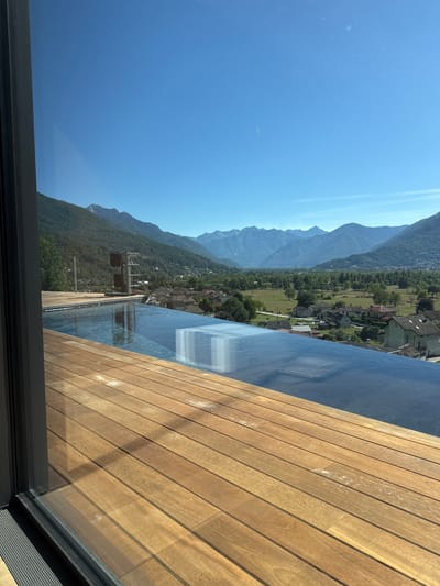 The media file shows a view from a rooftop pool overlooking a valley in Masera, Italy. The pool's edge is in the foreground, with a wooden deck on the right. The water in the pool reflects the clear blue sky and the surrounding mountains.
The valley below is filled with lush green trees and dotted with houses, suggesting a rural or semi-rural setting. In the distance, majestic mountains rise, their peaks softened by the haze of a sunny day. The sun is high in the sky, indicating it is likely midday. The overall atmosphere is one of tranquility and natural beauty. There are no people visible in the image.