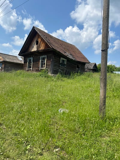 An eye-level, medium shot captures a weathered, dilapidated wooden house on a bright, sunny day in Новомуллакаево, Russia. The house, constructed from dark logs, features a rusted tin roof with visible holes and damage, and the wooden siding appears aged and worn. Two windows on the visible front facade are broken, with panes missing or cracked, and the frames are in disrepair. The house is surrounded by tall, overgrown green grass, indicative of a rural or abandoned setting. A wooden utility pole with dark electrical wires is positioned to the right of the frame, its surface weathered and marked. In the foreground, a plastic bottle lies on the grass, contributing to a sense of neglect. A second, similar wooden house is partially visible to the left of the main subject, and another smaller structure is seen in the background behind the primary house. The sky above is a vibrant blue, dotted with fluffy white clouds.