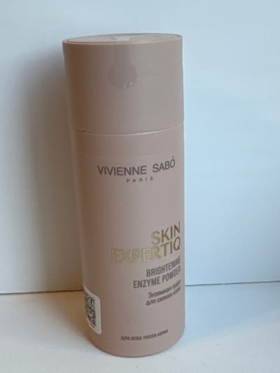 The media file displays a close-up, side-view of a cylindrical cosmetic product, specifically a "Vivienne Sabo Skin Expertiq Brightening Enzyme Powder." The product is a light, neutral-toned tube, lying horizontally on a white surface, likely a tabletop. The lighting suggests an indoor environment, possibly a domestic setting, with no distinct time of day or weather cues.
The tube features prominent gold lettering. The brand name "VIVIENNE SABO" and "PARIS" are clearly visible. The product name "SKIN EXPERTIQ" is also legible, followed by "BRIGHTENING ENZYME POWDER." Below this, in smaller text, is "Энзимная пруд" and "DRA CARINA" which appear to be in Cyrillic script, possibly indicating the product's intended use or ingredients. A QR code is partially visible on the lower right side of the tube. A faint, circular smudge is present on the upper left portion of the tube. The background is predominantly a soft, light blue, suggesting a wall or a backdrop behind the product. The overall impression is a simple, clean presentation of a beauty product. The context provided by "Яромінскі сельскі Савет, Belarus" indicates the product is being showcased or photographed within this specific region of Belarus.