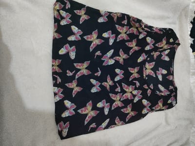 In Juja ward, Kenya, a black garment patterned with multicolored butterflies lies on a light-colored, textured surface. The butterflies feature shades of pink, yellow, blue, and white. The garment appears to be an article of clothing such as a top or dress, positioned vertically within the frame. No individuals, actions, or interactions are present in the scene. The background surface is uniform in color and texture.