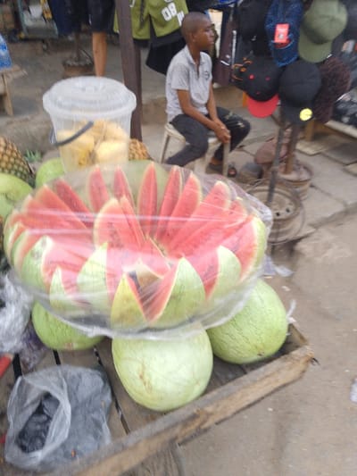A street vendor's stand displays watermelons, some pre-cut into red wedges and wrapped in plastic. A bucket of sliced pineapple sits nearby. Behind the fruit, a young boy sits on a small stool, seemingly waiting for customers. Clothes and hats hang in the background, suggesting a varied market scene. The stand itself appears to be constructed of rough wooden planks.