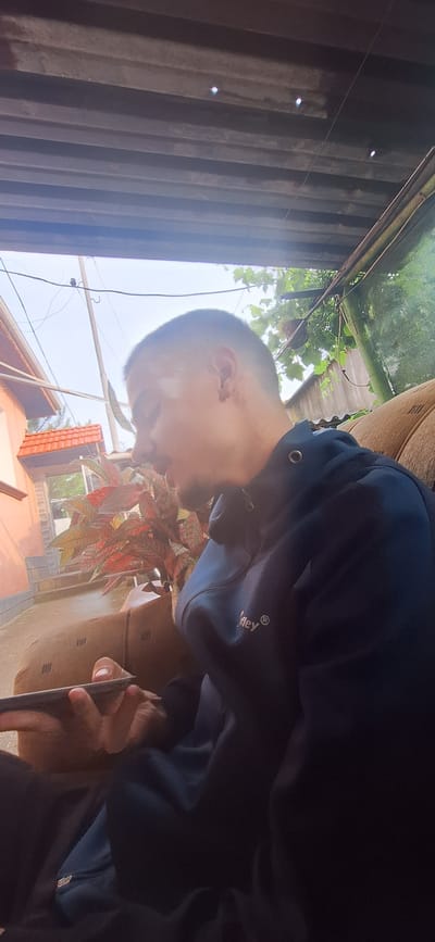 A young man is sitting on a sofa outdoors, holding a mobile phone to his ear. He is wearing a dark blue hooded jacket with a small logo on the chest. The background shows a building with a tiled roof, some foliage, and a corrugated metal roof overhead. The overall scene suggests a casual, possibly residential, outdoor setting. The location in the city of Morava, Bulgaria is not evident from the image itself.