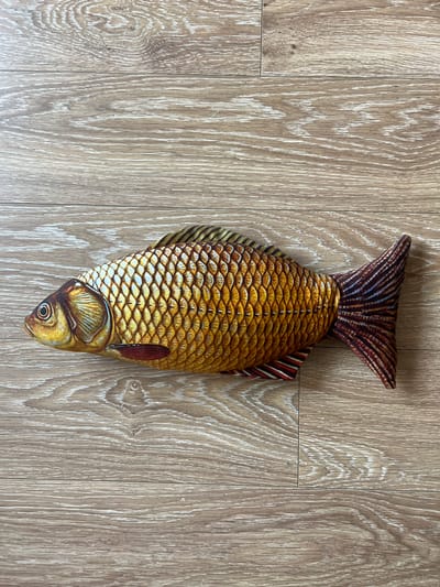 This image shows a close-up, top-down view of a plush toy fish lying on a light brown wooden floor. The fish is positioned vertically in the center of the frame, with its head pointing downwards. The floor is made of planks with a visible wood grain pattern, featuring a mix of light and darker brown tones, and some white accents that highlight the texture.
The fish toy is detailed and appears to be made of fabric. It has a realistic design, with scales depicted in various shades of yellow and gold, edged with darker lines. The tail fin is broad and fan-shaped, with alternating stripes of dark brown and reddish-brown, interspersed with thin blue and dark red lines. The body of the fish also has a darker stripe running down its length, mimicking a lateral line. The head of the fish is rendered with attention to detail, including a visible eye with a black pupil and a reddish-brown gill cover. A smaller, reddish-brown pectoral fin is visible on the right side of the fish's body. The overall impression of the fish is that of a carp or a similar freshwater species.
The lighting suggests an indoor environment, likely illuminated by artificial light or ambient daylight, as there are no strong shadows indicative of direct sunlight. The setting appears to be a room with a wooden floor. No text is visible in the image.