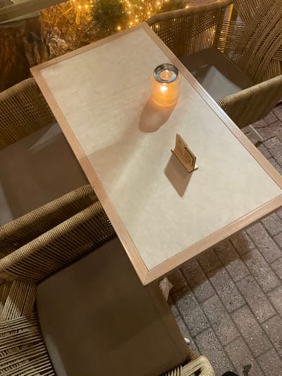 The image captures an outdoor table setting at night in Chaidari, Greece, viewed from an elevated angle. A rectangular table with a light, perhaps off-white, surface and a warm wooden frame dominates the center. On the table, a clear glass jar containing an illuminated object, likely a candle or small lamp, casts a soft, warm glow and a noticeable shadow. Beside it rests a small, dark wooden stand or holder, angled slightly. Text is visible on this stand, but it is largely illegible due to resolution, although phrases such as "Thank you for..." can be vaguely discerned on the upper part.
Surrounding the table are chairs with woven, rope-like backs and seats, some appearing to have dark brown cushions. The background on the left features more of this woven material and a blurred display of bright, small lights, reminiscent of a festive or decorative tree, suggesting an inviting atmosphere. To the right, the ground is paved with rectangular bricks, indicating a walkway or patio area.
The time of day is clearly night, as evidenced by the artificial lighting from the table lamp and decorative lights, and the overall low ambient light. There are no people visible, but the setup implies a relaxed, comfortable, and potentially social dining or lounge setting. The warm lighting contributes to a cozy and inviting mood.
