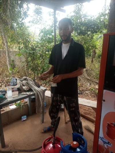 An adult male stands centrally within an outdoor LPG refilling station in Omicha Umu Awada, Nigeria. He possesses a dark beard and wears a black collared shirt over a white undershirt, dark patterned trousers, and blue sandals. The man faces the camera directly, holding bundles of Nigerian Naira currency in both hands, appearing to count them. To his left, a grey plastic table supports a clear plastic bottle with a handwritten label, a bundle of light-colored fabric, a calculator, a dark-colored smartphone, and other assorted papers and a black plastic bag. On the red-brown dirt ground, near the man's feet, are a red LPG cylinder and a blue LPG cylinder, both fitted with valves and connected by a black hose, indicative of a gas dispensing operation. A red-orange gas dispensing unit is partially visible on the far right. A small broom lies on the ground to the man's right. The background features dense green vegetation, including trees and bushes, a section of a concrete wall, and a chain-link fence. A rudimentary wooden bench is present behind the man. The scene is illuminated by natural daylight, casting soft shadows.