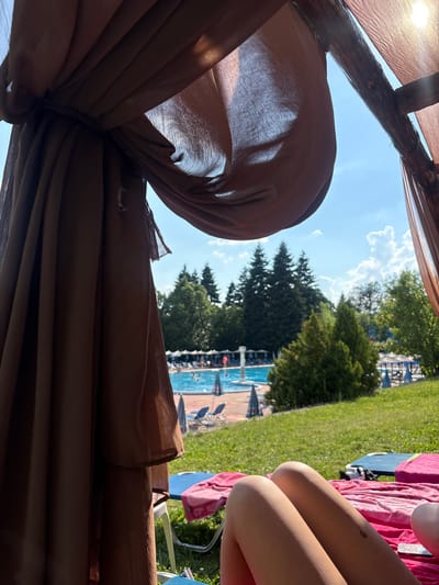 In Rudartsi, Bulgaria, a person's legs are visible in the foreground, resting on a pink towel on a lounge chair. The individual is positioned within a shaded cabana, indicated by the brown fabric drapes in the upper left and right portions of the frame. Beyond the cabana, a swimming pool area is visible, with people swimming and sunbathing. Lounge chairs and umbrellas are set up around the pool, and a grassy area with trees and foliage surrounds the pool and lounge area. The sky is blue with scattered white clouds, suggesting a sunny day.